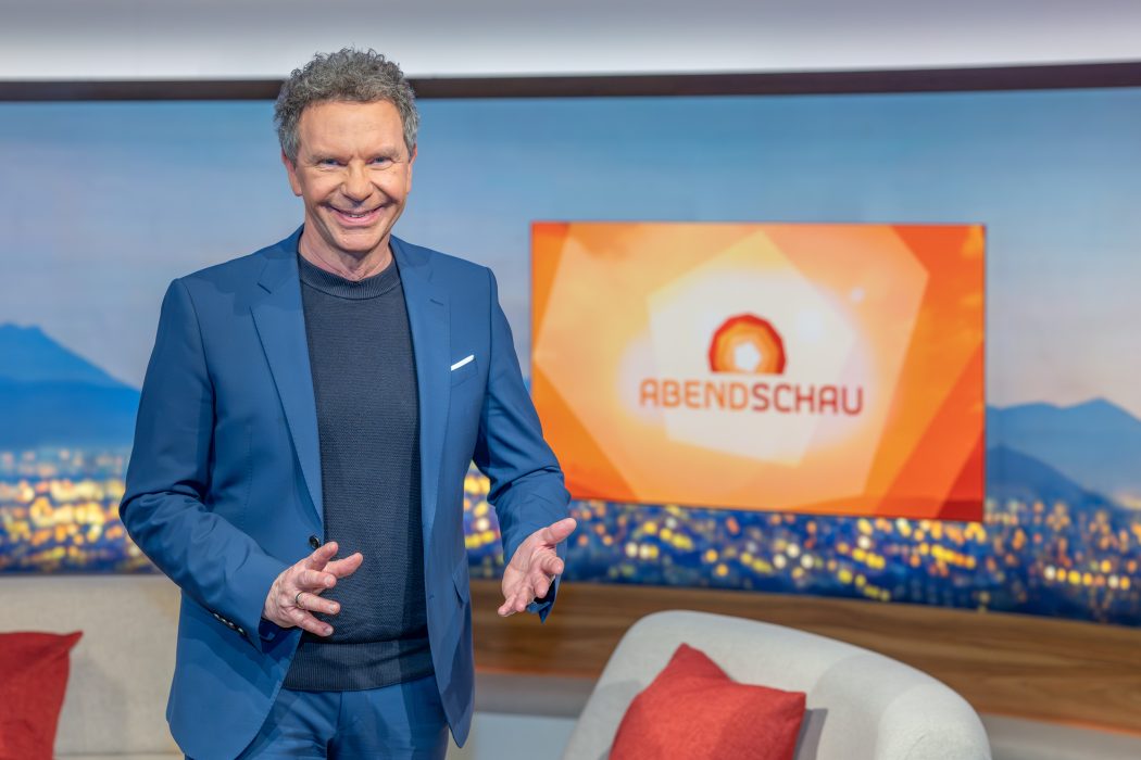 Roman Röll im blauen Anzug vor dem Abendschau-Bildschirm im BR-Studio